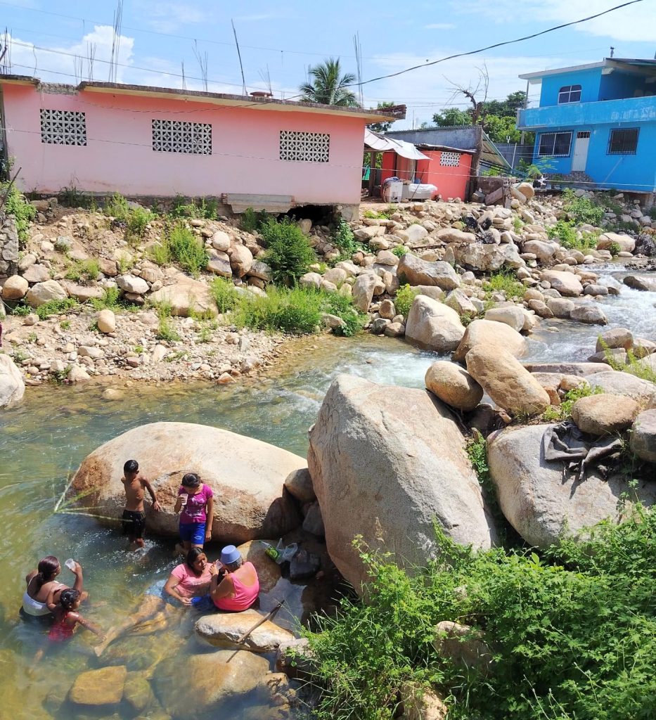 Photo shows a family of 5 people in a river. Two women sitting facing each other, one in the water and one on a rock. Two children stand in the water leaning against a large rock. Another woman and smaller child face them. The woman is holding a cup and the child is leaning on her arm. There are colourful buildings in the background.