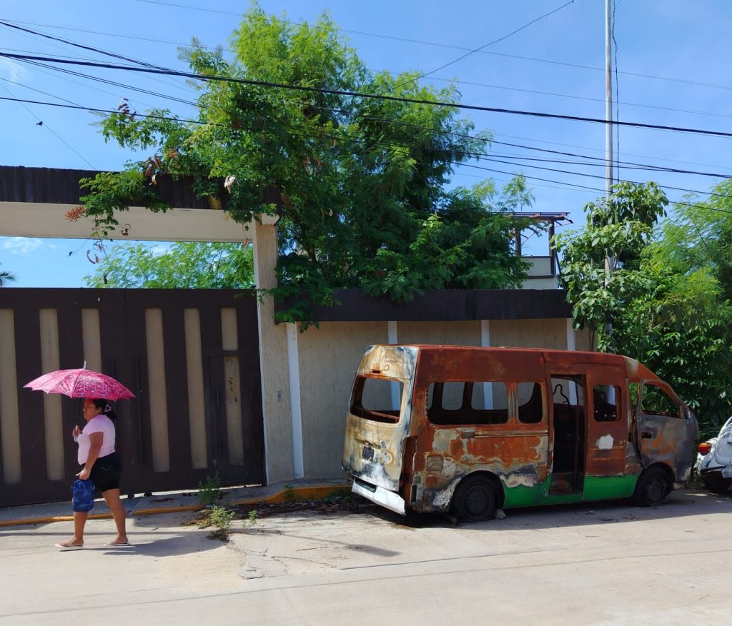 The photo shows an abandoned yellow van with no door in the street with no other traffic. A woman holding a pink umbrella is walking past.